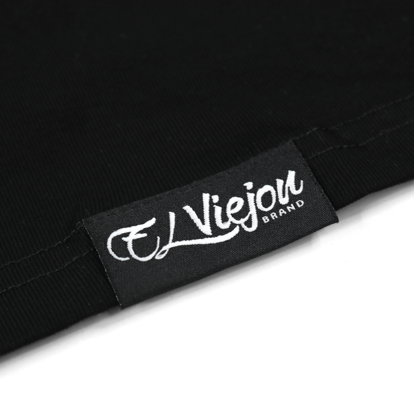 Close-up of a black fabric tag with "El Viejon Brand" in white script, sewn onto a black polo shirt.