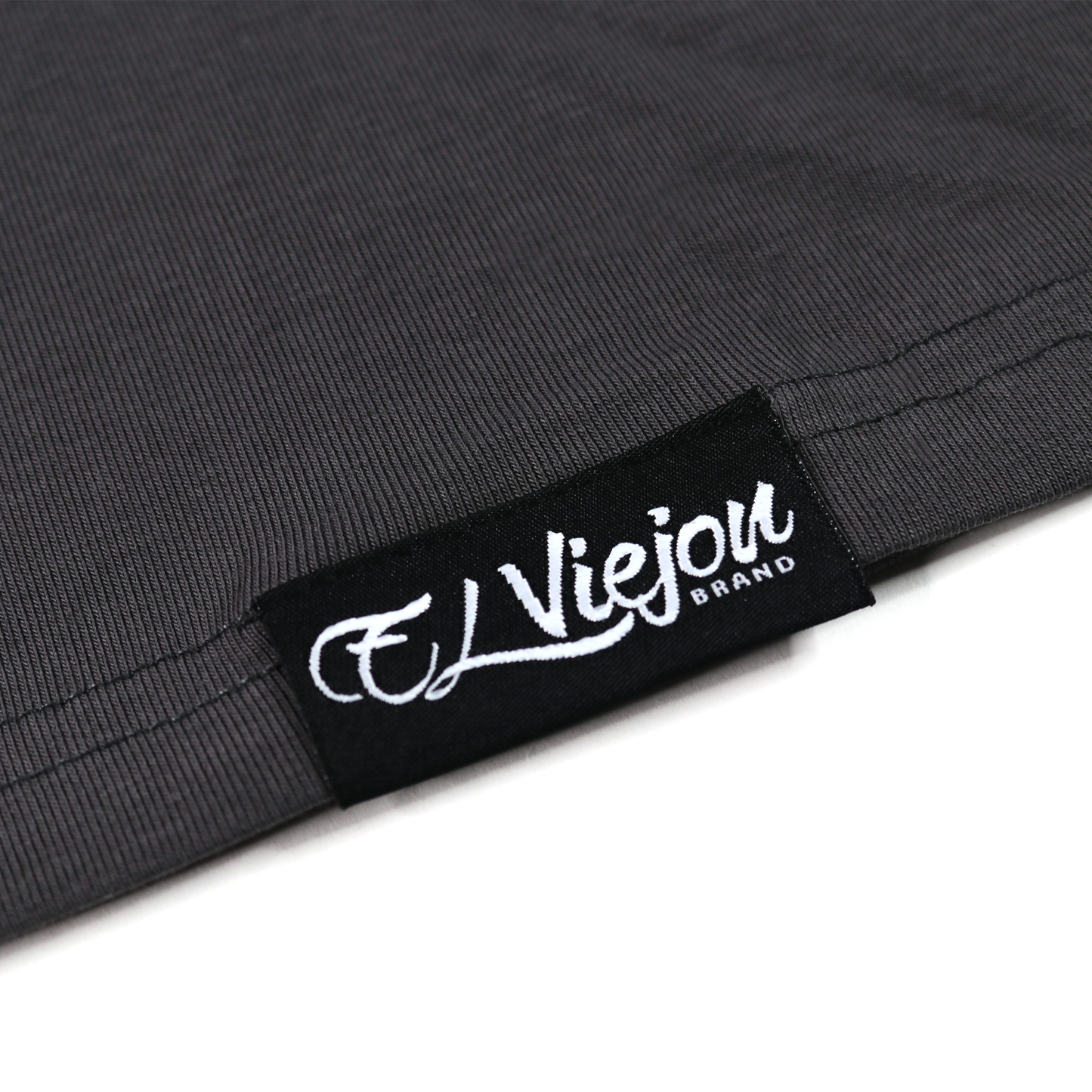 Close-up of a charcoal polo shirt hem with a black "El Viejon Brand" tag in white script.